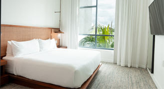 The image shows a modern bed with pillows and a view outside of the flat.