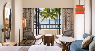 Room with bed, two chairs and wooden table, and a view of the ocean from the balcony