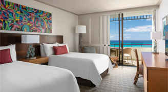 A view of a room with two beds, a desk and a view of the ocean in the background.