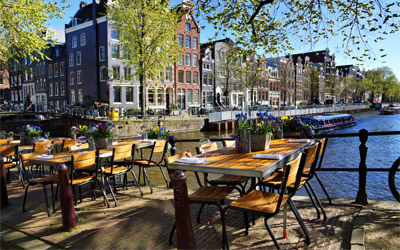 Restaurant tables lining the beautiful canals of Amsterdam under blue skies.